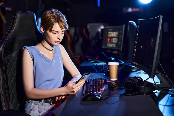 Kurzhaarige Frau, die ihr Smartphone in der Nähe des Computers auf dem Schreibtisch benutzt, Cybersport-Spielerin im Computerclub — Stockfoto