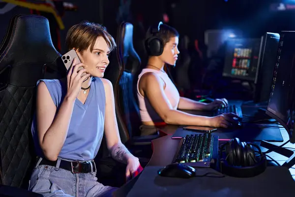 Mulher sentada em uma mesa de computador falando por telefone perto de amigo jogando no jogo de computador no clube — Fotografia de Stock