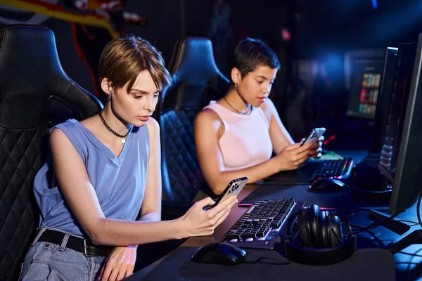 Two young women sit at computer desk while scrolling their phones in cybersport game club — Stock Photo