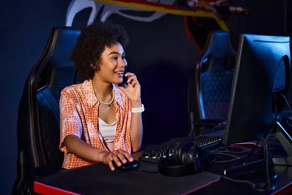 Mujer negra feliz se sienta en su computadora y tiene llamada telefónica cerca de la computadora, club de cybersport - foto de stock