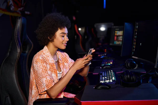 Feliz mujer negra se sienta cerca de la computadora, su rostro iluminado por la pantalla azul del teléfono inteligente - foto de stock