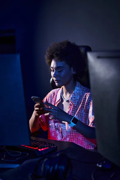 Афроамериканська жінка з кучерявим волоссям у синьому світлі від монітора комп'ютера, використовуючи свій смартфон — стокове фото