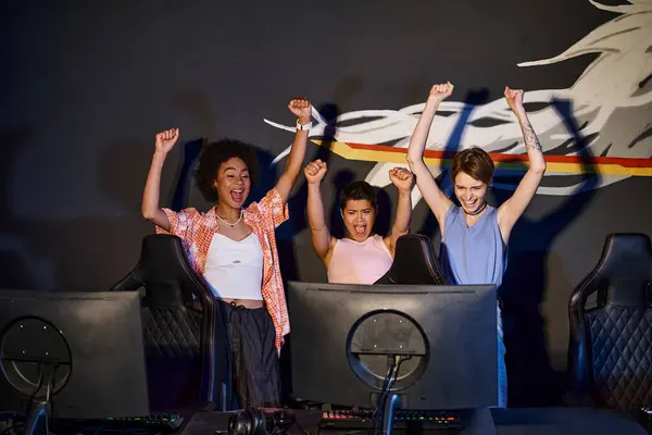 Multicultural happy women looking at computer monitor and celebrating victory, raised hands — Stock Photo