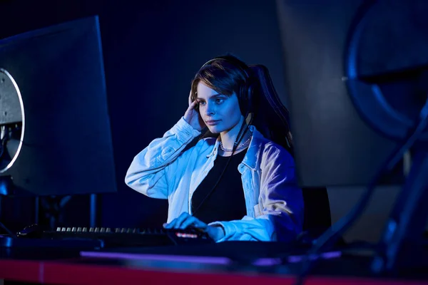 Young woman wearing headphones and looking at computer in a blue-lit room, cybersport game concept — Stock Photo