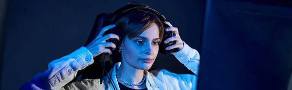 Focused woman wearing headphones and looking at computer in a blue-lit room, cybersport game banner — Stock Photo
