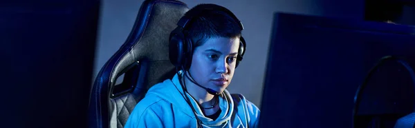 Gamer enfocado con pelo corto mirando a la computadora en una habitación con luz azul, jugador con sudadera con capucha - foto de stock