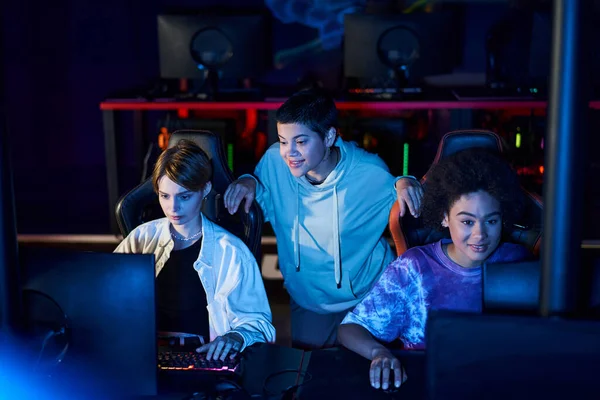 Diversas mujeres que participan en juegos de ciberdeporte, utilizando computadoras y sonriendo en la habitación con luz azul - foto de stock