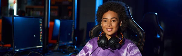 Happy african american female gamer with headphones sitting on comfortable gaming chair, banner — Stock Photo