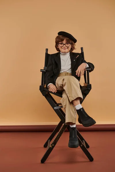 Felice ragazzo in abbigliamento casual si siede sulla sedia regista su sfondo beige, bambino e professione — Foto stock