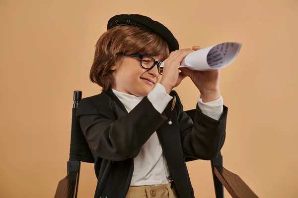 Boy dressed in stylish clothing sits on director chair and looking through hole in rolled paper — Stock Photo