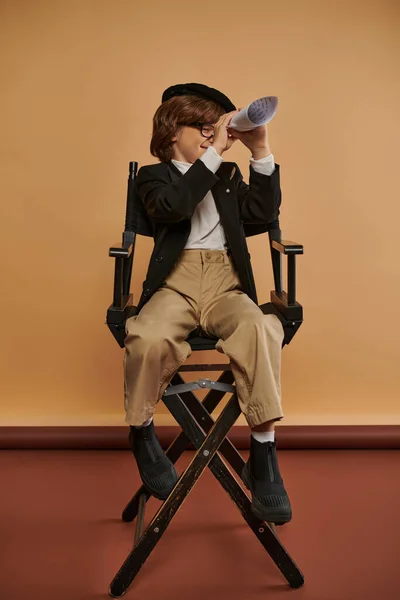 Carino ragazzo in abbigliamento elegante si siede sulla sedia regista e guardando attraverso il buco in carta arrotolata — Foto stock
