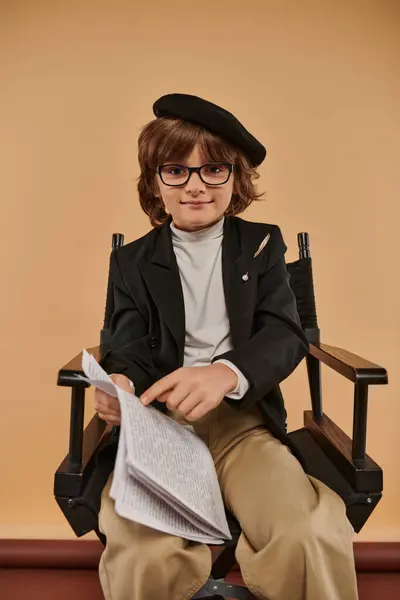 Niño en boina y gafas se sienta en silla de director, señalando con el dedo en el papel, profesión y niño - foto de stock