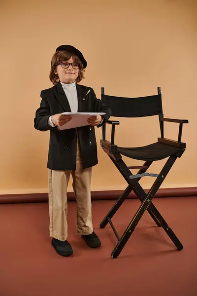 Glücklicher Junge steht stolz neben dem Regiestuhl, in Jacke und Hose gekleidet, mit Papieren in der Hand — Stockfoto
