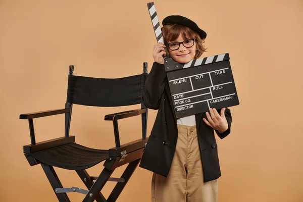 Cineasta niño en ropa de moda posa felizmente con un clapperboard en la mano contra la pared beige - foto de stock