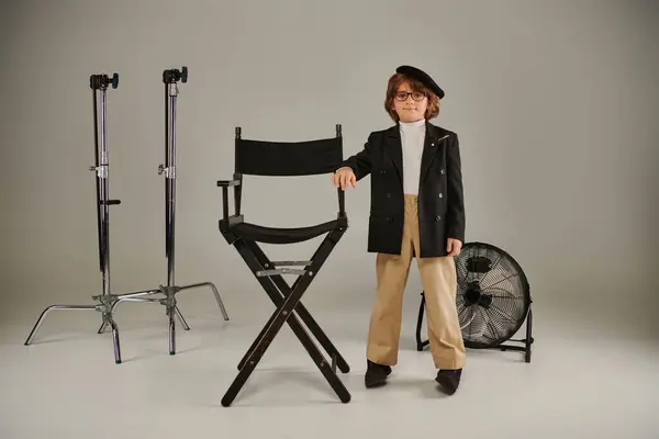 Netter Junge in Baskenmütze und schicker Freizeitkleidung steht selbstbewusst neben dem Regiestuhl auf grauem Hintergrund — Stockfoto