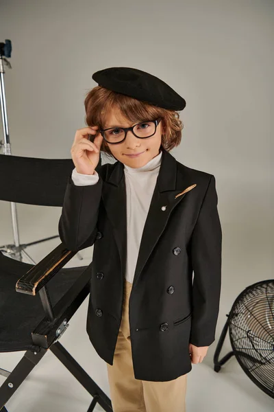 Стильный мальчик в куртке и берете, улыбаясь на сером фоне, ребенок в качестве режиссера — стоковое фото