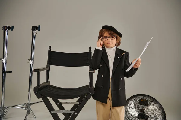 Стильный мальчик в пиджаке и берет с собой бумагу со сценарием на сером, ребенок в качестве режиссера — стоковое фото