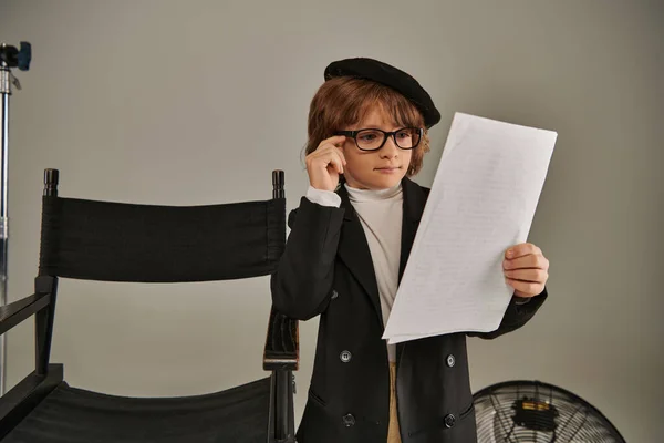 Chico con estilo en gafas y boina guion de lectura en papel sobre gris, niño como director de cineasta - foto de stock