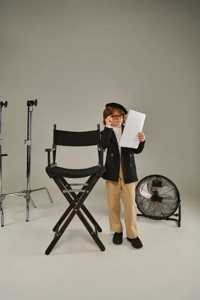 Stilvoller Junge mit Brille und Baskenmütze liest Drehbuch auf Papier auf Grau, Kind als Regisseur — Stockfoto