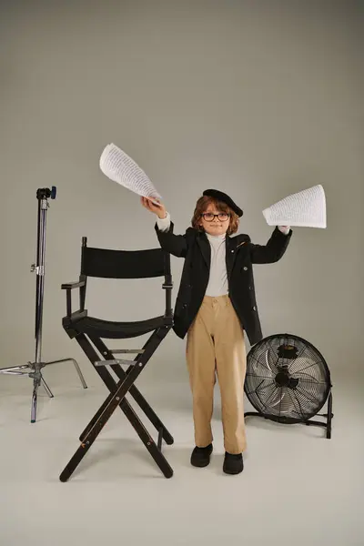 Menino elegante em óculos e boina segurando roteiro em papéis em cinza, criança como diretor do cineasta — Fotografia de Stock
