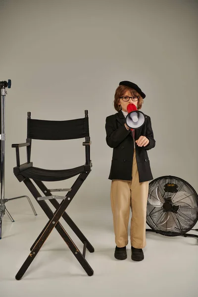 Stilvolles Kind steht selbstbewusst neben Regiestuhl und spricht in Megafon, grauem Hintergrund — Stockfoto