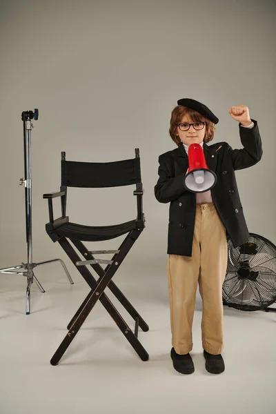 Um jovem ativista em boina está com megafone e cadeira de diretor em cinza, menino como cineasta — Fotografia de Stock