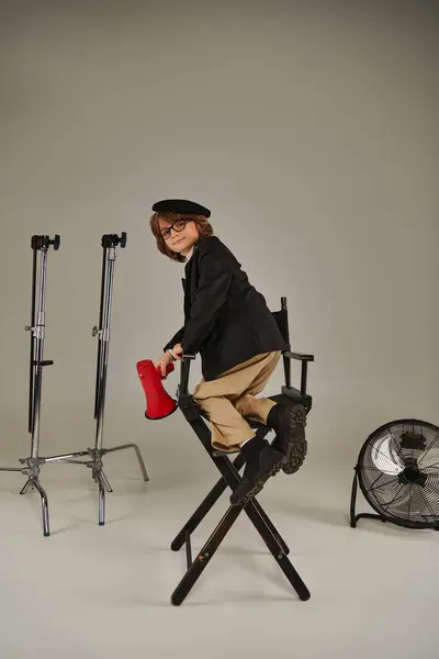 Мальчик-режиссер забирается на режиссерское кресло и держит красный мегафон в руке на сером фоне — стоковое фото
