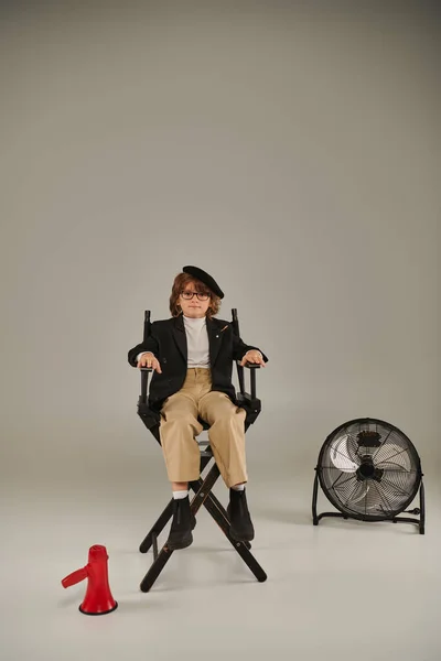 Seguro cineasta chico en boina y gafas sentado en director silla cerca de megáfono en gris - foto de stock