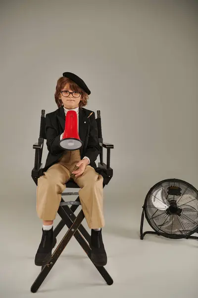 Confident filmmaker boy in beret and glasses sitting on director chair and holding red megaphone — Stock Photo
