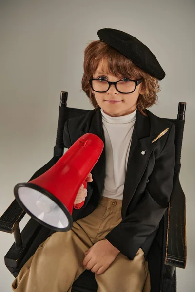 Lindo cineasta chico en boina y gafas sentado en la silla del director y la celebración de megáfono rojo - foto de stock