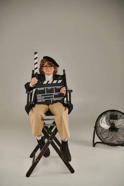 Niño en boina y gafas sosteniendo el tablero de aplausos como sentado en la silla del director, concepto de fabricación de películas - foto de stock