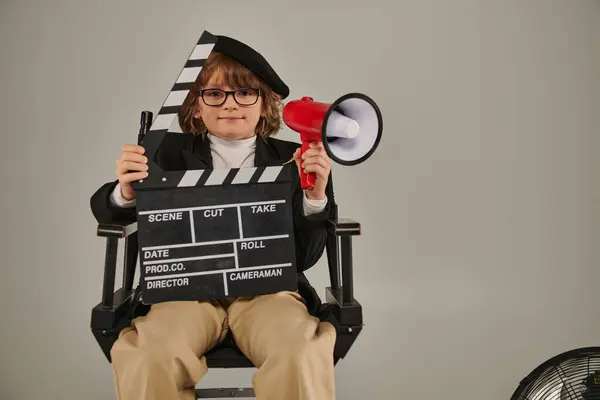 Мальчик в берете и очках, держащий доску объявлений и красный мегафон, сидящий на режиссерском стуле — стоковое фото