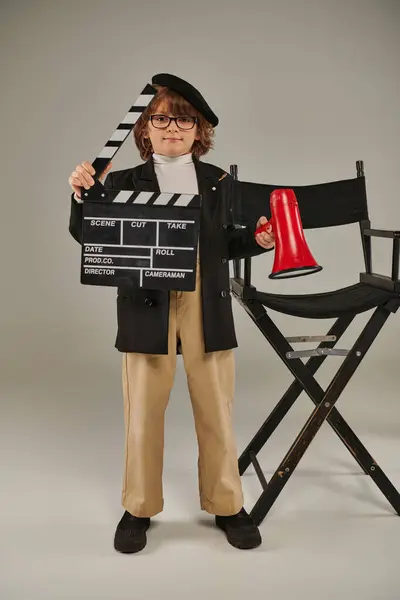 Мальчик в берете и очках, держащий доску хлопания и красный мегафон и стоящий рядом с режиссерским креслом — стоковое фото