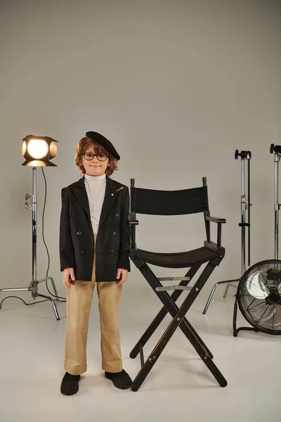 Хорошо одетый мальчик в берцах и очках, стоящий перед режиссерским креслом, трезвый на заднем плане — стоковое фото