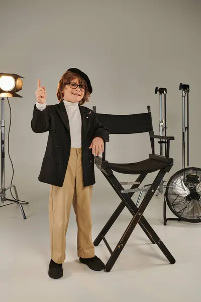 Feliz chico con estilo en boina y gafas de pie delante de la silla del director, softbot en el fondo - foto de stock