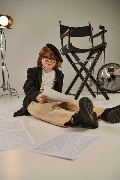 Niño en gafas y boina leyendo guion y sentado en el suelo, niño como director de cineasta - foto de stock