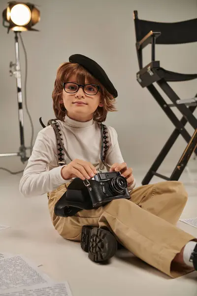 Niño en boina sosteniendo cámara retro y sentado en el suelo cerca de la silla de director, joven fotógrafo - foto de stock