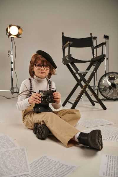 Niño en boina sosteniendo la cámara vintage y sentado en el suelo cerca de la silla de director, joven fotógrafo - foto de stock