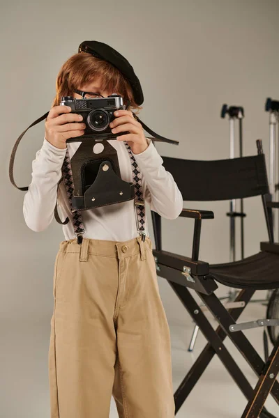 Ragazzo in berretto cattura il momento sulla fotocamera retrò vicino alla sedia del regista, giovane fotografo in studio — Foto stock