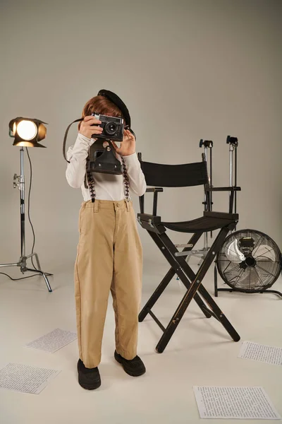Menino na boina captura o momento na câmera retro perto da cadeira do diretor, jovem fotógrafo em estúdio — Fotografia de Stock