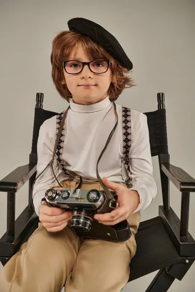Niño en boina y gafas sosteniendo la cámara retro mientras está sentado en la silla del director, joven fotógrafo - foto de stock