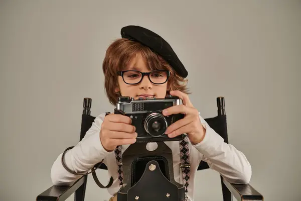 Niño en boina y gafas que sostienen la cámara retro mientras está sentado en la silla del director, joven fotógrafo - foto de stock