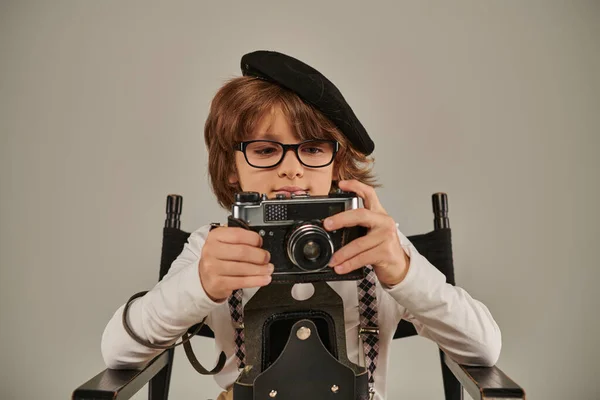 Kid in beret and glasses holding retro camera while sitting on director chair, young photographer — Stock Photo