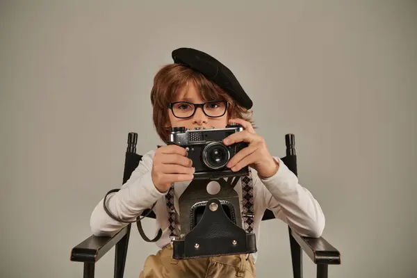 Boy in beret and glasses holding vintage camera while sitting on director chair, young photographer — Stock Photo