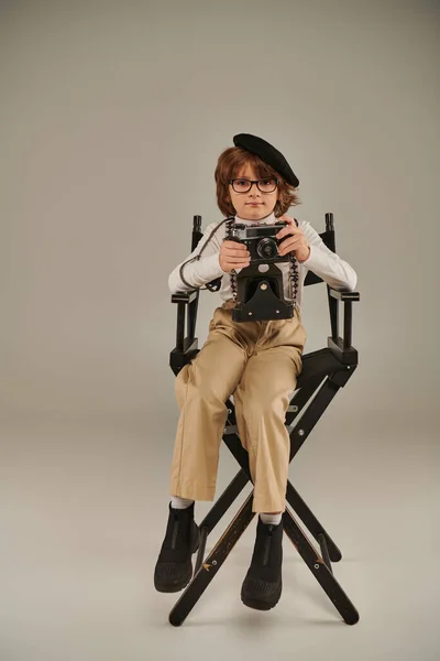 Giovane fotografo in berretto e occhiali con fotocamera retrò seduto sulla sedia del regista — Foto stock