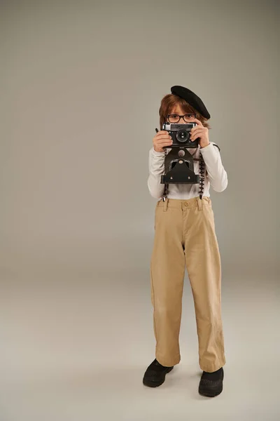 Young photographer in beret and suspenders holding retro camera while standing in studio — Stock Photo