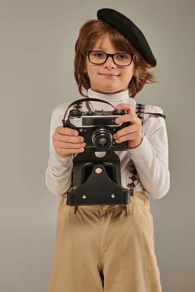 Happy boy, young photographer in beret and suspenders holding retro camera while standing in studio — Stock Photo