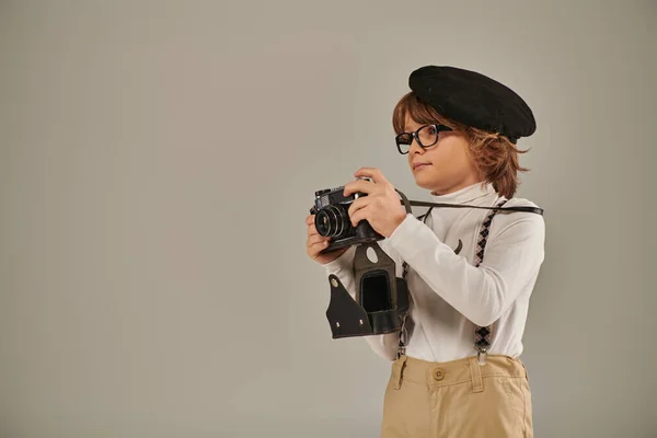Cute boy, young photographer in beret and suspenders taking photo on retro camera in studio — Stock Photo