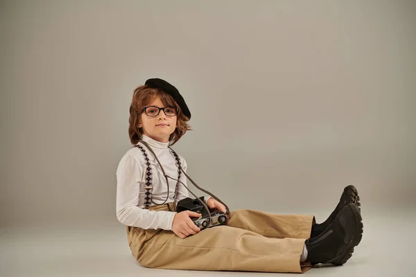 Jeune photographe en béret et lunettes tenant un appareil photo rétro et assis sur le sol, enfant en jarretelles — Photo de stock