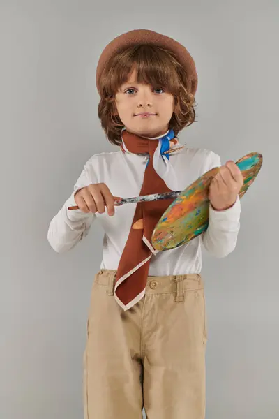 Young artist holding paint brush and palette, cute kid in beret and scarf standing on grey backdrop — Stock Photo